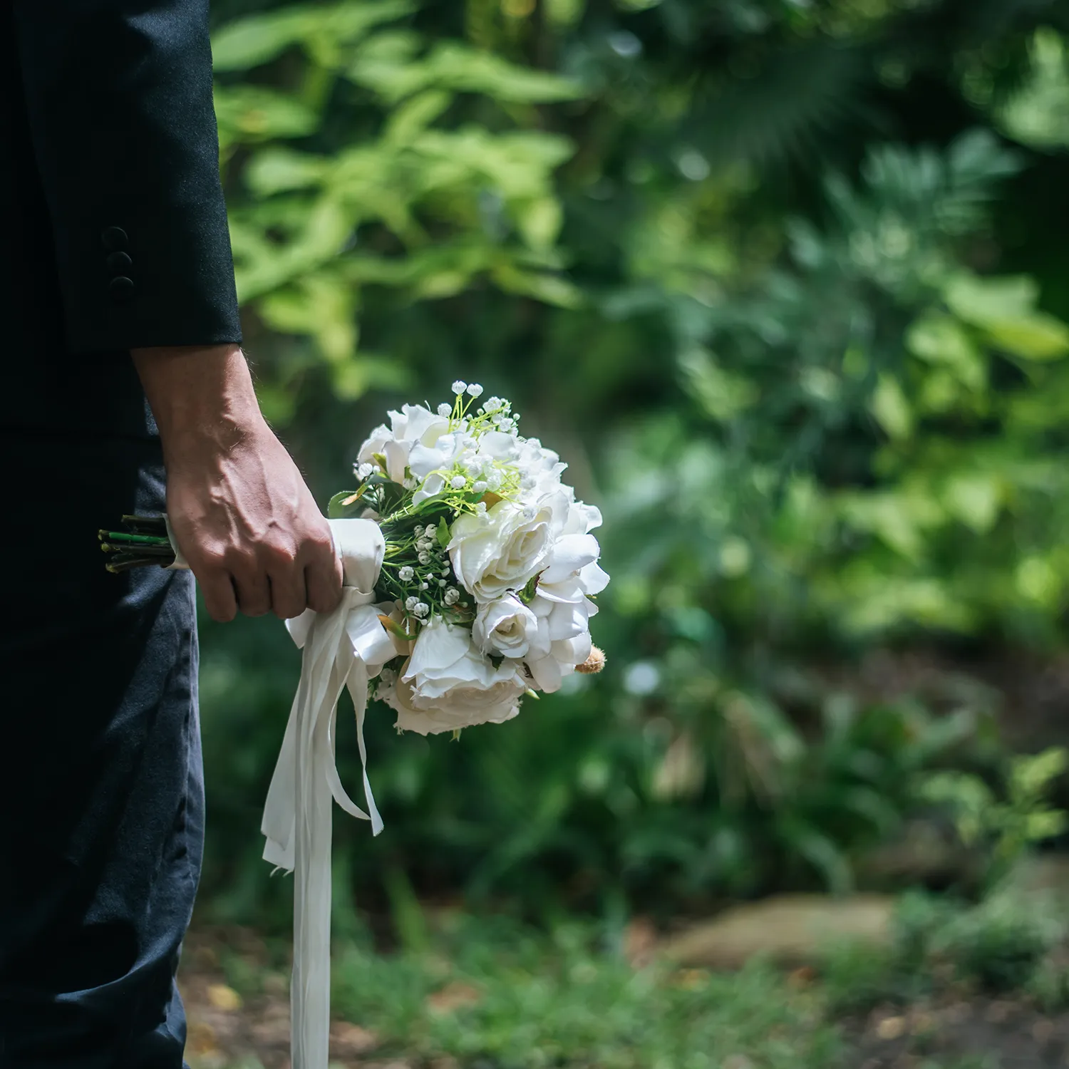 Trauergast im dunklen Anzug hält einen weißen Rosenstrauß mit Schleife im Grünen
